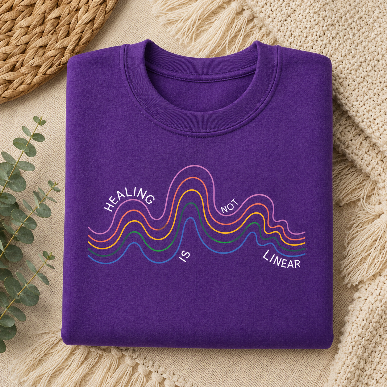 A folded purple crewneck sweatshirt with a colorful graphic reading 'HEALING IS NOT LINEAR' in white text over wavy rainbow lines, styled on a beige textured blanket with a woven mat and eucalyptus leaves.