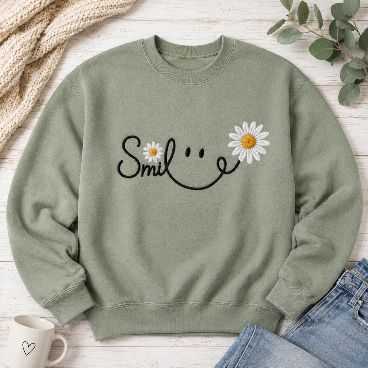 An olive green crewneck sweatshirt with a black smiley face graphic and white daisy flowers, styled in a flat lay on a white wood background with a mug, jeans, and eucalyptus.