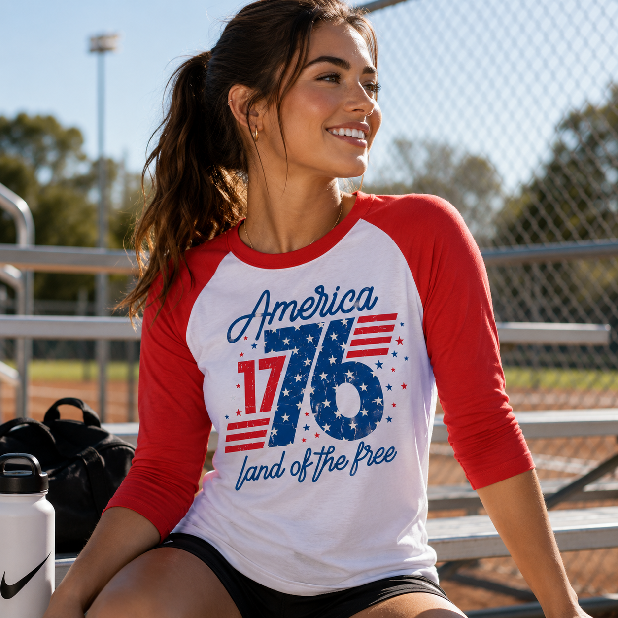 Woman wearing a red and white baseball shirt with 'America 1776 land of the free' design, sitting outdoors.