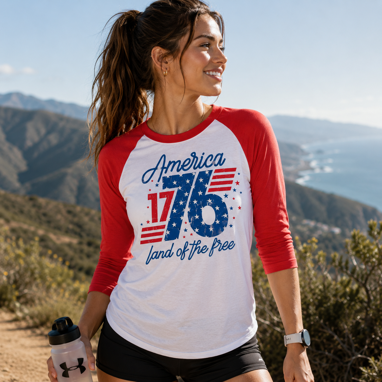 Woman wearing a red and white baseball shirt with 'America 1776 land of the free' text, standing outdoors with mountains in the background.