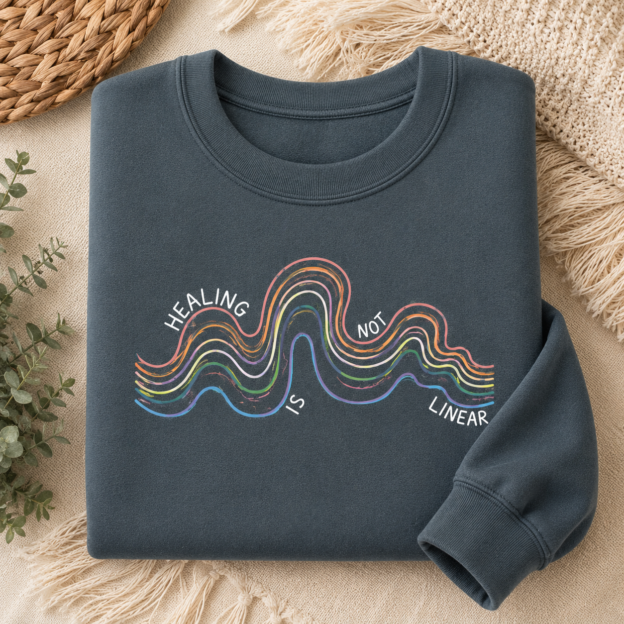A folded dark slate blue crewneck sweatshirt with a colorful graphic of wavy lines and the text 'HEALING IS NOT LINEAR' printed on the front, styled on a beige textured blanket with a woven basket and green leaves.
