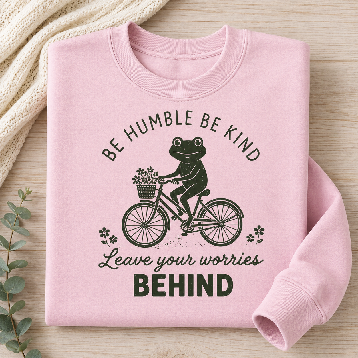 A folded light pink crewneck sweatshirt with a black graphic print of a frog riding a bicycle and the text 'BE HUMBLE BE KIND Leave your worries BEHIND'. The sweatshirt is styled on a light wood background with a cream knit blanket and eucalyptus leaves.