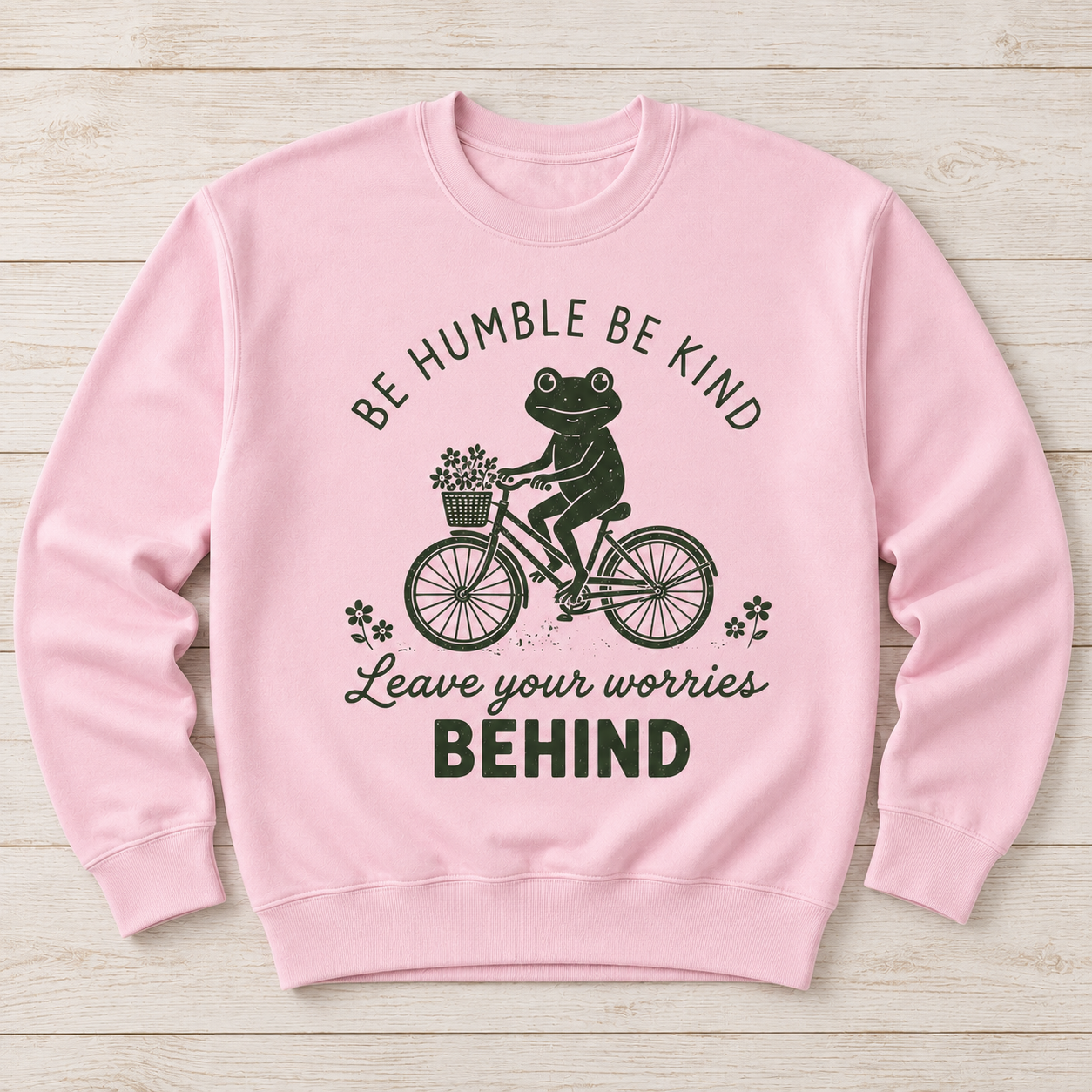 A light pink crewneck sweatshirt with a dark green graphic of a frog riding a bicycle and the text 'BE HUMBLE BE KIND Leave your worries BEHIND', displayed flat on a light wood background.