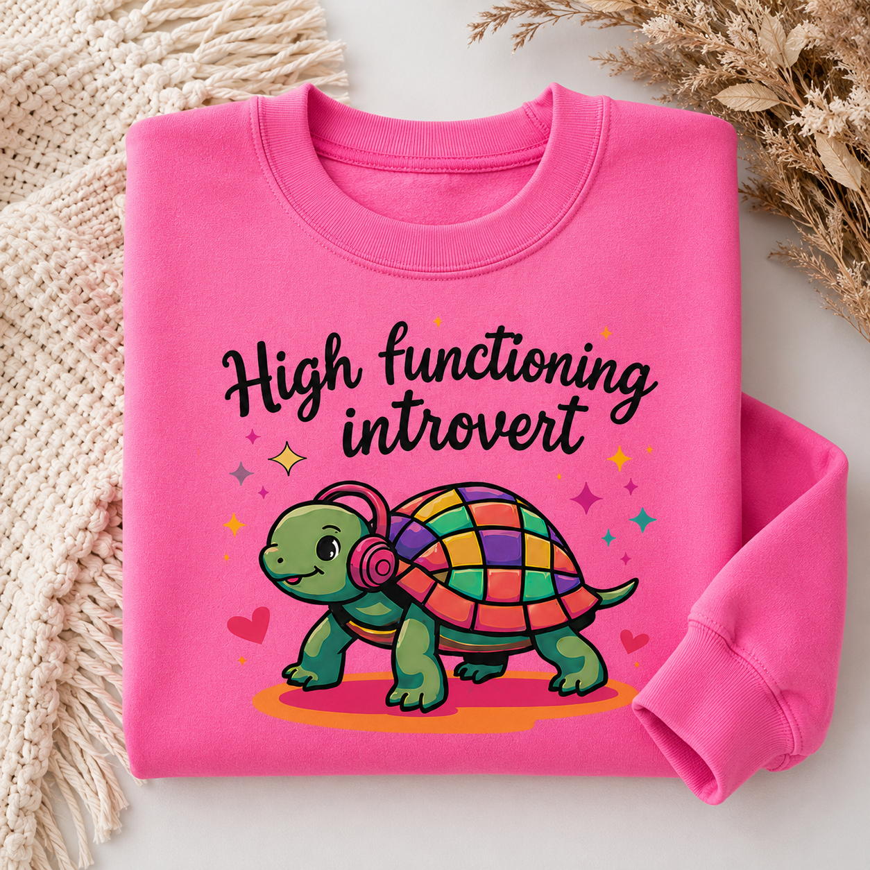 A folded bright pink sweatshirt on a light surface with decorative knit fabric and dried flowers. The sweatshirt has a colorful cartoon turtle wearing headphones with the text 'High functioning introvert' in black script.