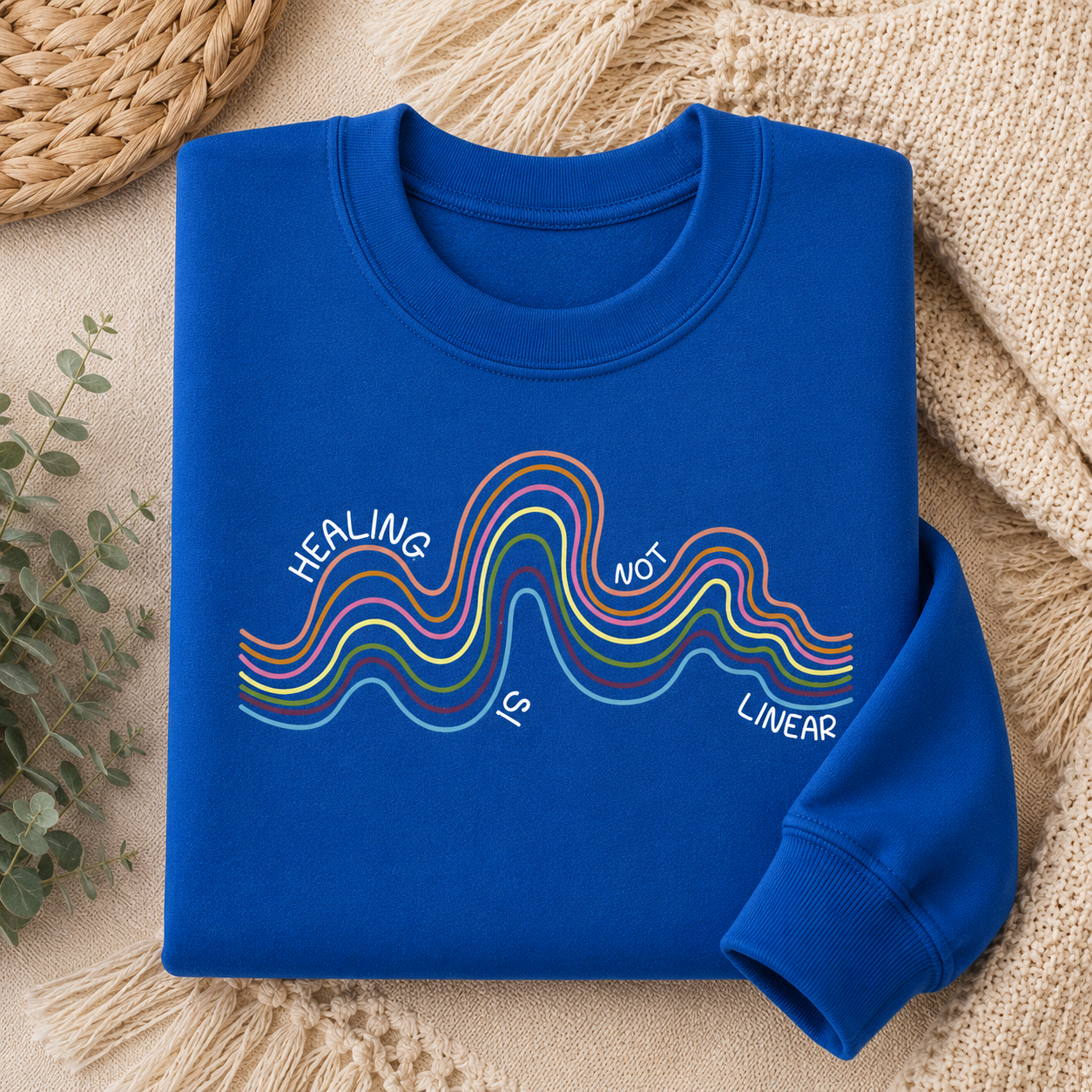 A folded royal blue crewneck sweatshirt with a colorful wavy graphic and the text 'HEALING IS NOT LINEAR' printed on the front, styled on a beige knit blanket with a woven mat and eucalyptus leaves.
