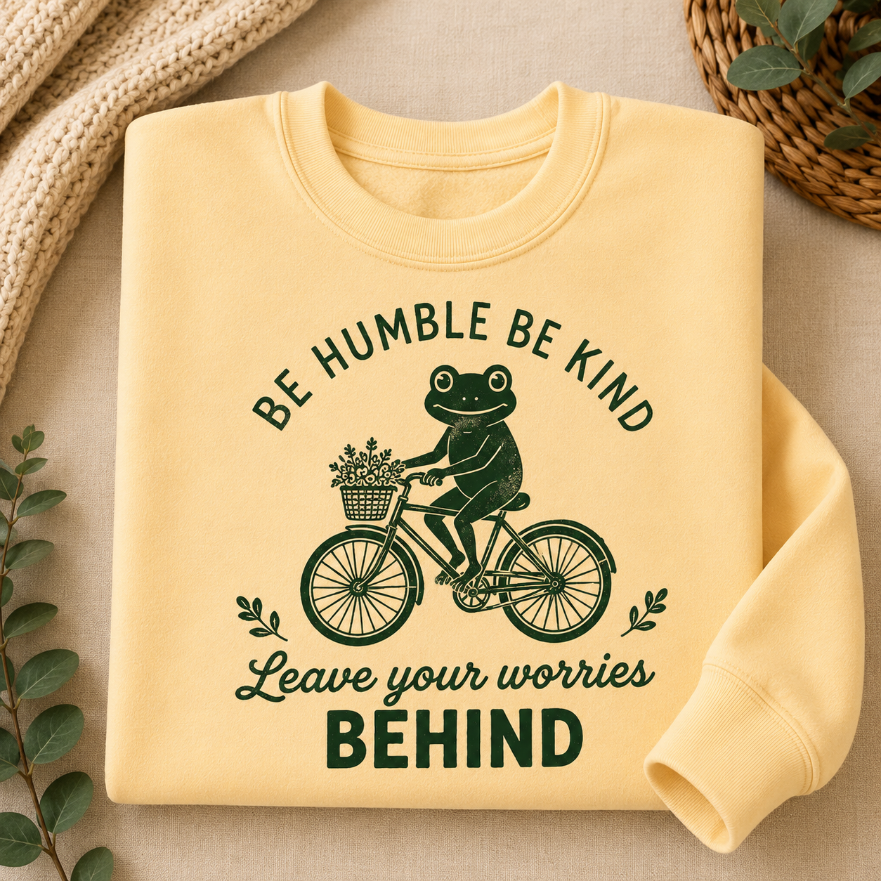 A folded pale yellow sweatshirt with a dark green graphic of a frog riding a bicycle and the text 'BE HUMBLE BE KIND Leave your worries BEHIND', styled on a beige fabric background with decorative leaves and a knitted textile.