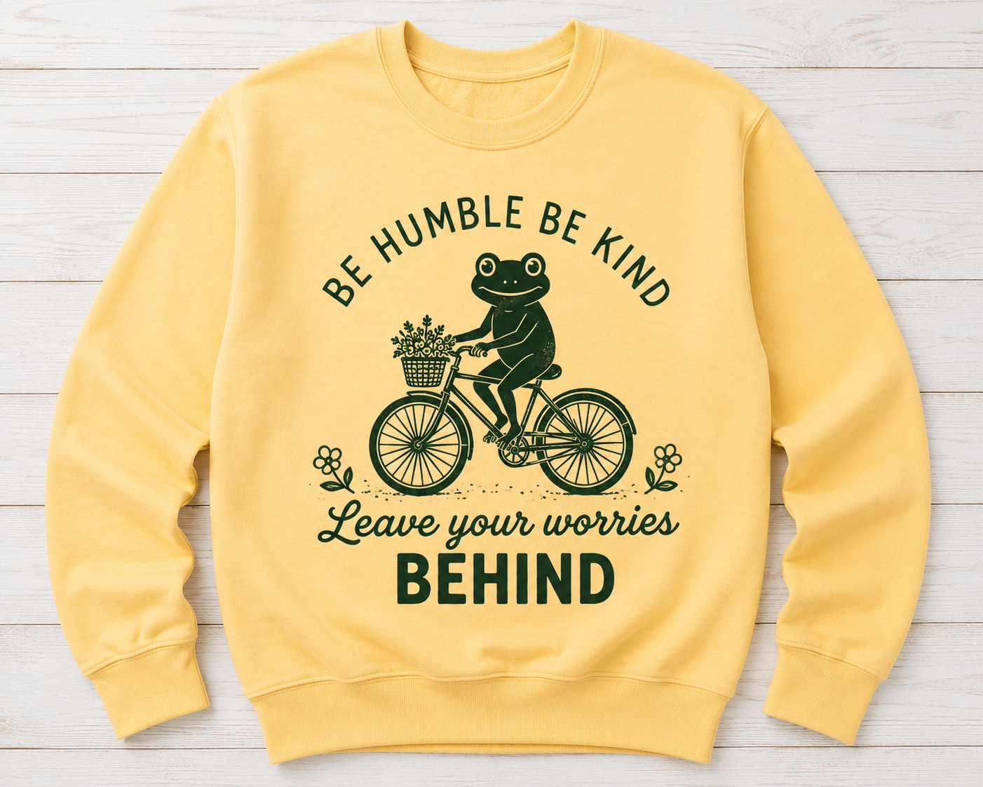 A pale yellow crewneck sweatshirt laid flat on a white wood plank background. The front features a dark green graphic of a frog riding a bicycle with flowers in the basket, surrounded by the text 'BE HUMBLE BE KIND Leave your worries BEHIND'.