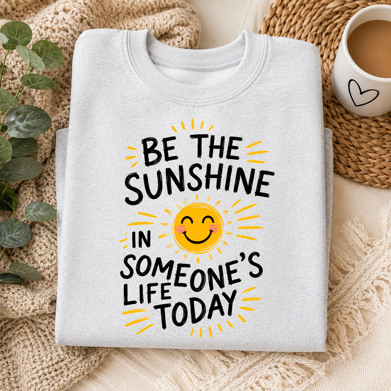 A folded light gray sweatshirt with a graphic print that reads 'BE THE SUNSHINE IN SOMEONE'S LIFE TODAY' in black text around a yellow smiling sun, styled on a beige knit blanket with a cup of coffee and green leaves nearby.