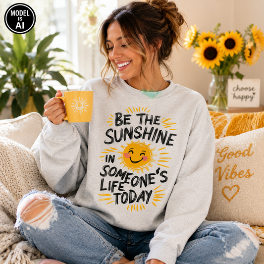 A woman sitting on a couch in a cozy living room setting, wearing a light gray sweatshirt with a yellow and black graphic that reads 'BE THE SUNSHINE IN SOMEONE'S LIFE TODAY' with a smiling sun face. She is holding a yellow mug with a sun design.