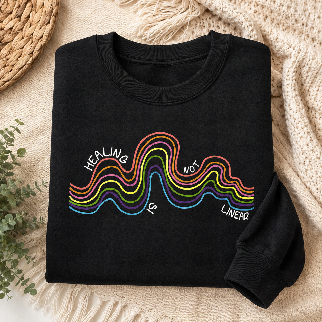 A folded black long-sleeve crewneck sweatshirt with a colorful wavy graphic and the text 'HEALING IS NOT LINEAR' printed on the front, styled on a beige knit blanket with a woven mat and greenery in the background.