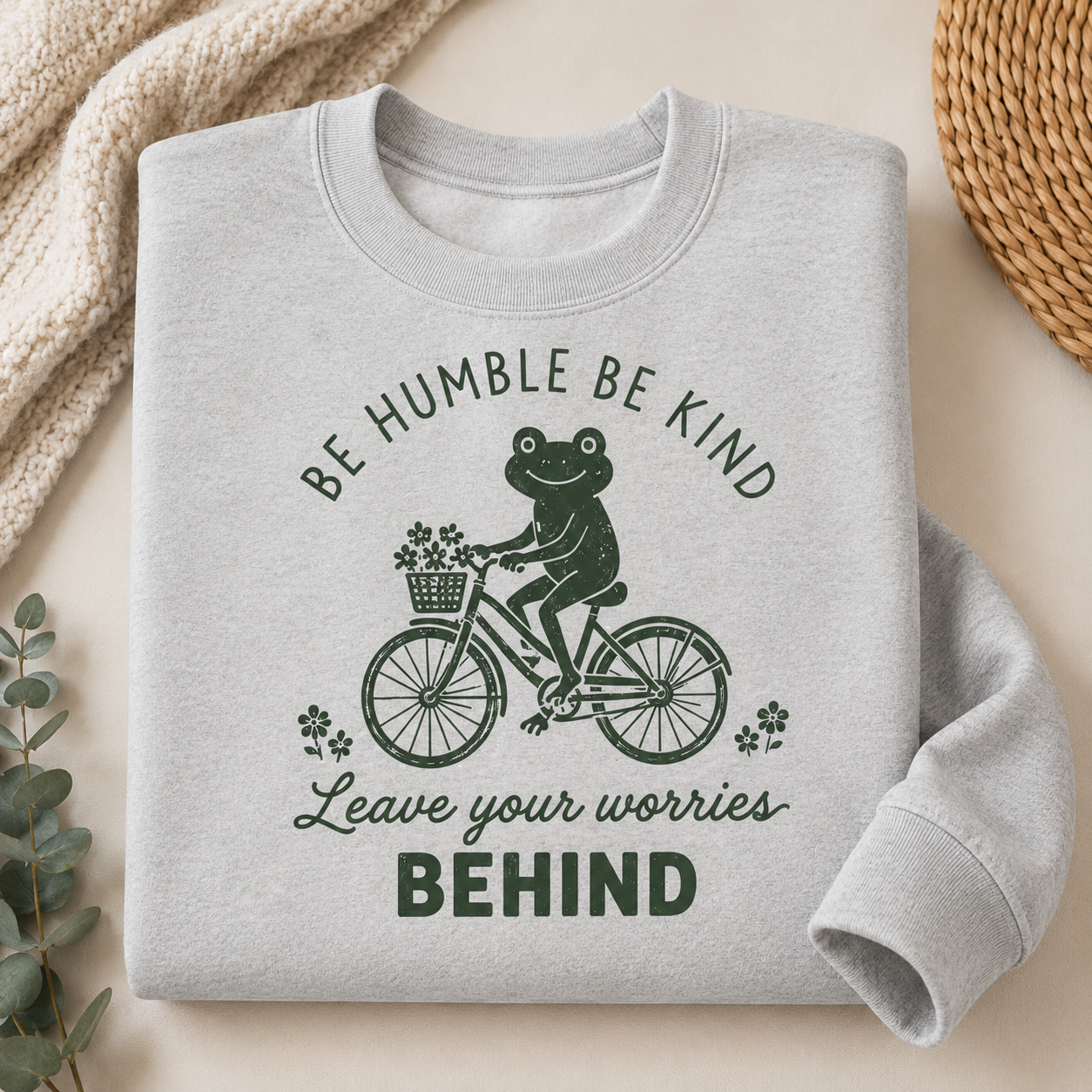 A light gray crewneck sweatshirt with a dark green graphic of a frog riding a bicycle and the text 'BE HUMBLE BE KIND Leave your worries BEHIND', styled in a flat-lay with a cream knit blanket, eucalyptus leaves, and a woven mat.