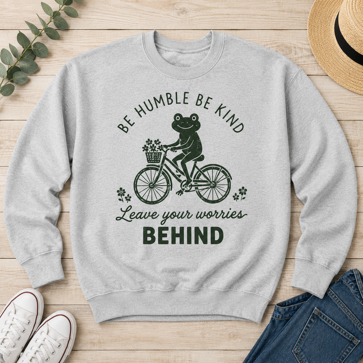 A light gray crewneck sweatshirt with a dark green graphic of a frog riding a bicycle and the text 'BE HUMBLE BE KIND Leave your worries BEHIND', laid flat on a light wood background with white sneakers, blue jeans, a straw hat, and eucalyptus leaves as styling props.