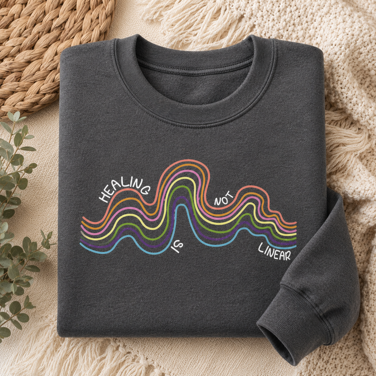 A dark charcoal gray crewneck sweatshirt with a colorful graphic of wavy lines and the text 'HEALING IS NOT LINEAR' printed on the front, styled flat on a cozy beige knit blanket with a woven mat and eucalyptus leaves.