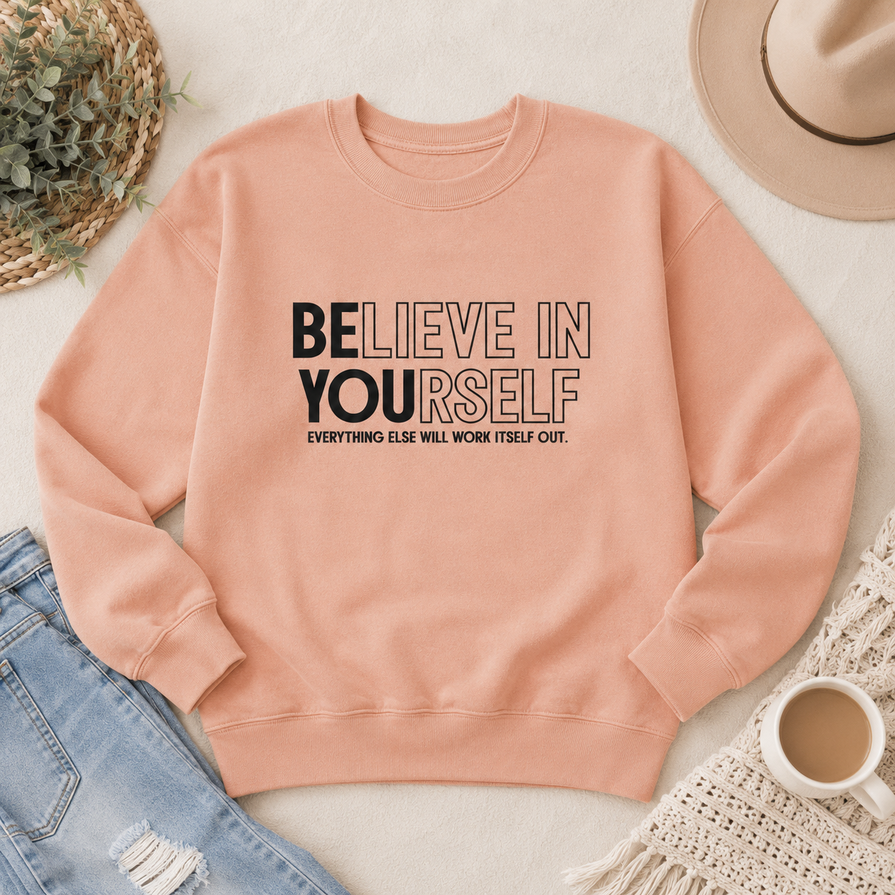 A peach-colored crewneck sweatshirt with the text 'BELIEVE IN YOURSELF' and 'EVERYTHING ELSE WILL WORK ITSELF OUT.' printed on the front.