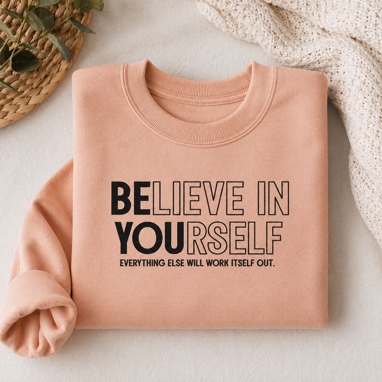A peach-colored crewneck sweatshirt with the motivational text 'BELIEVE IN YOURSELF' and 'EVERYTHING ELSE WILL WORK ITSELF OUT.' printed on the front, styled in a flat-lay arrangement with a woven basket, green leaves, and a cream knit blanket.