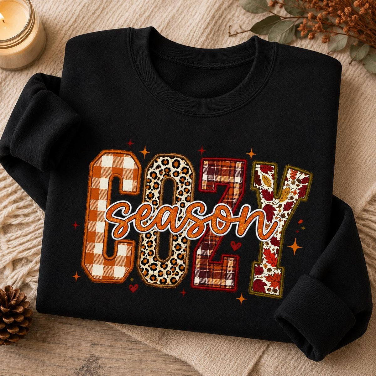 Folded black Cozy Season Sweatshirt with colorful patchwork fall graphic in a cozy autumn setting with candle and fall accents