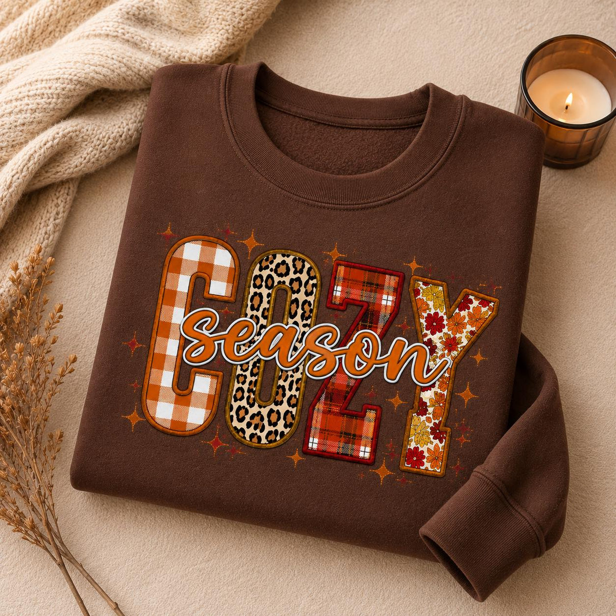 Folded Cocoa Cozy Season Sweatshirt with colorful patchwork fall graphic in a cozy autumn setting with candle and fall accents