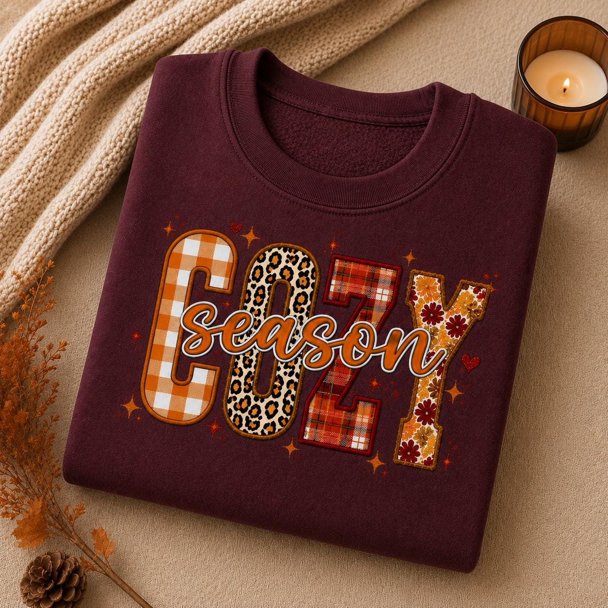 Folded Maroon Cozy Season Sweatshirt with colorful patchwork fall graphic in a cozy autumn setting with candle and fall accents