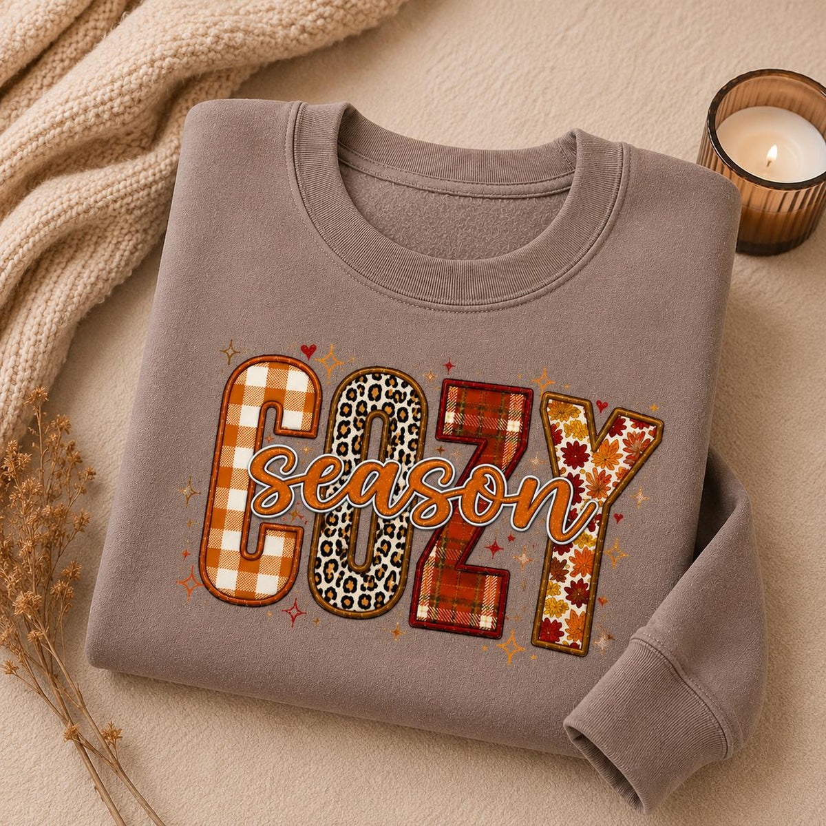 Folded Brown Cozy Season Sweatshirt with colorful patchwork fall graphic in a cozy autumn setting with candle and fall accents