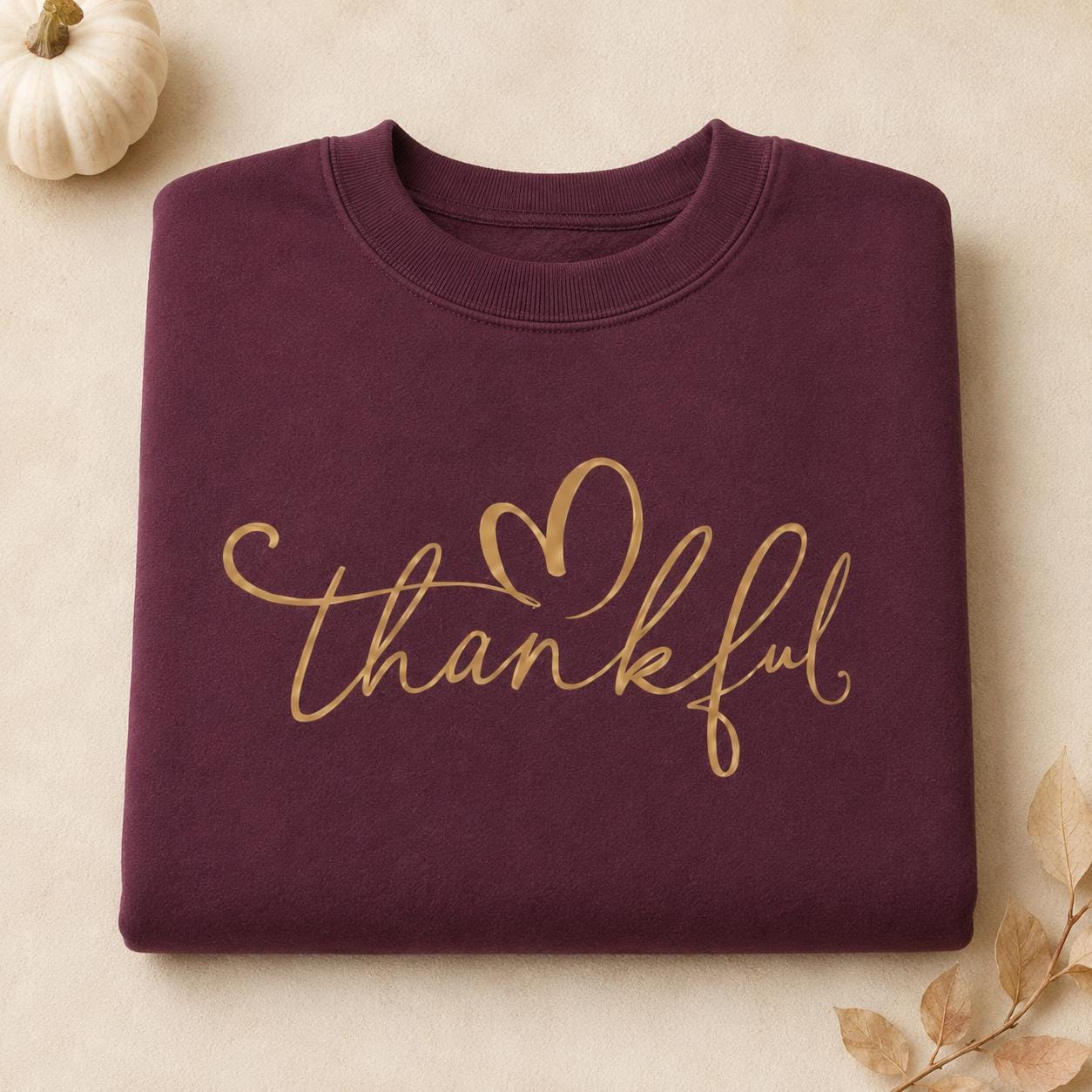 Folded maroon Thankful Sweatshirt with gold script heart design and fall pumpkin accents
