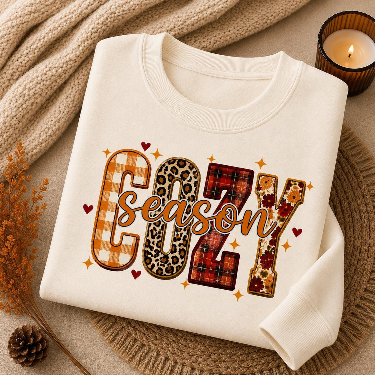 Folded Off White Cozy Season Sweatshirt with colorful patchwork fall graphic in a cozy autumn setting with candle and fall accents