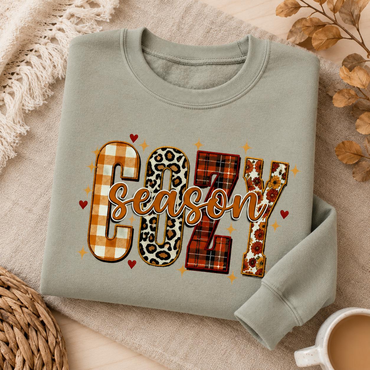 Folded Sage Green  Cozy Season Sweatshirt with colorful patchwork fall graphic in a cozy autumn setting with candle and fall accents