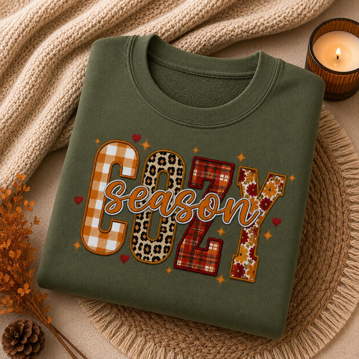 Folded Military Green Cozy Season Sweatshirt with colorful patchwork fall graphic in a cozy autumn setting with candle and fall accents