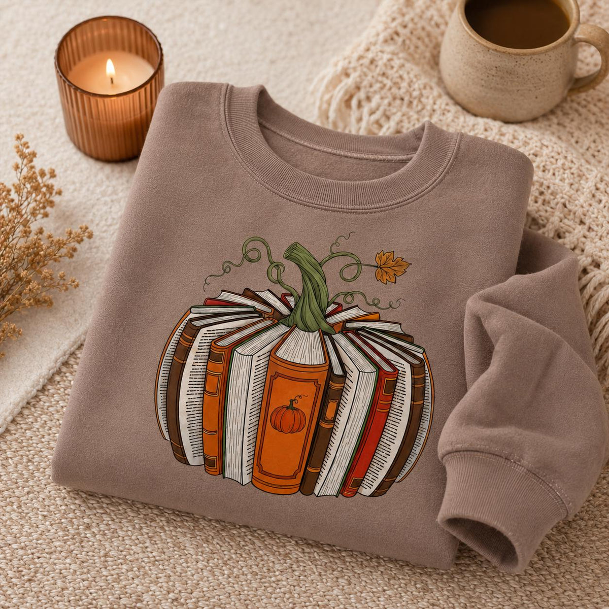 Folded brown Fall Book Lover Sweatshirt with pumpkin-shaped book graphic in a cozy autumn setting with coffee and candle
