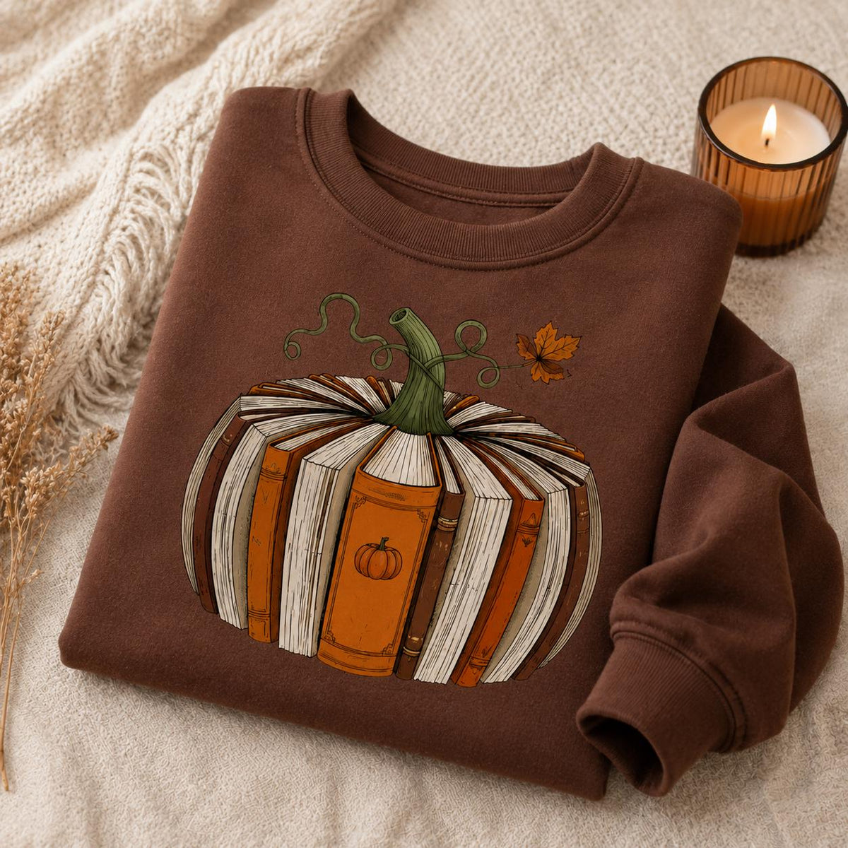 Folded brown Fall Book Lover Sweatshirt with pumpkin-shaped book graphic in a cozy autumn setting with candle and knit blanket
