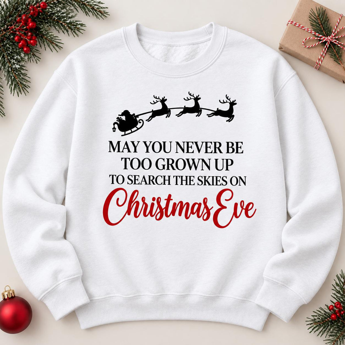 White Santa sleigh Christmas Eve sweatshirt laid flat with holiday greenery.