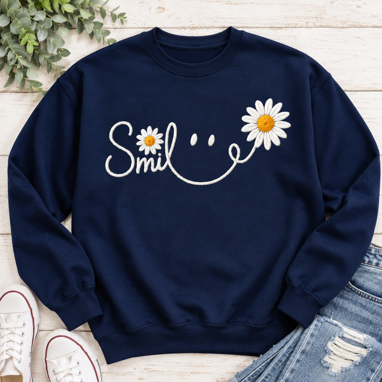 A navy blue crewneck sweatshirt with a white embroidered smiley face made from the word 'Smile' and two daisy flowers, styled in a flat lay with distressed blue jeans, white sneakers, and green leaves on a light wood background.