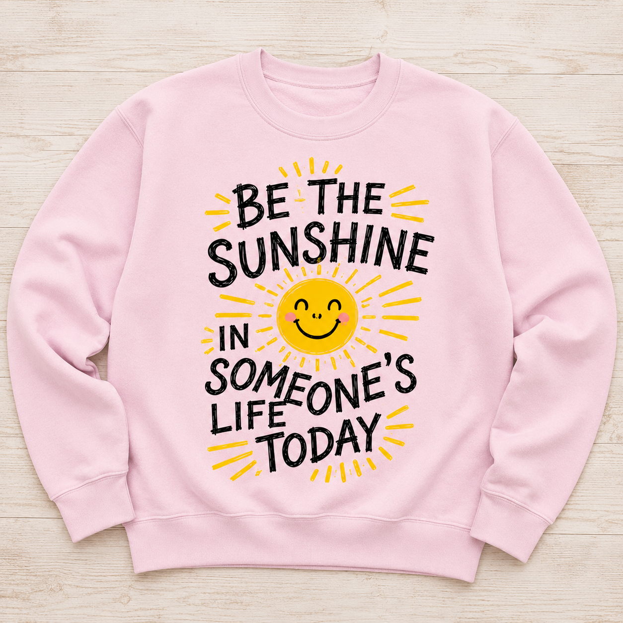 A light pink crewneck sweatshirt laid flat on a light wood background, featuring a graphic design with the text 'BE THE SUNSHINE IN SOMEONE'S LIFE TODAY' in black and yellow around a smiling yellow sun.