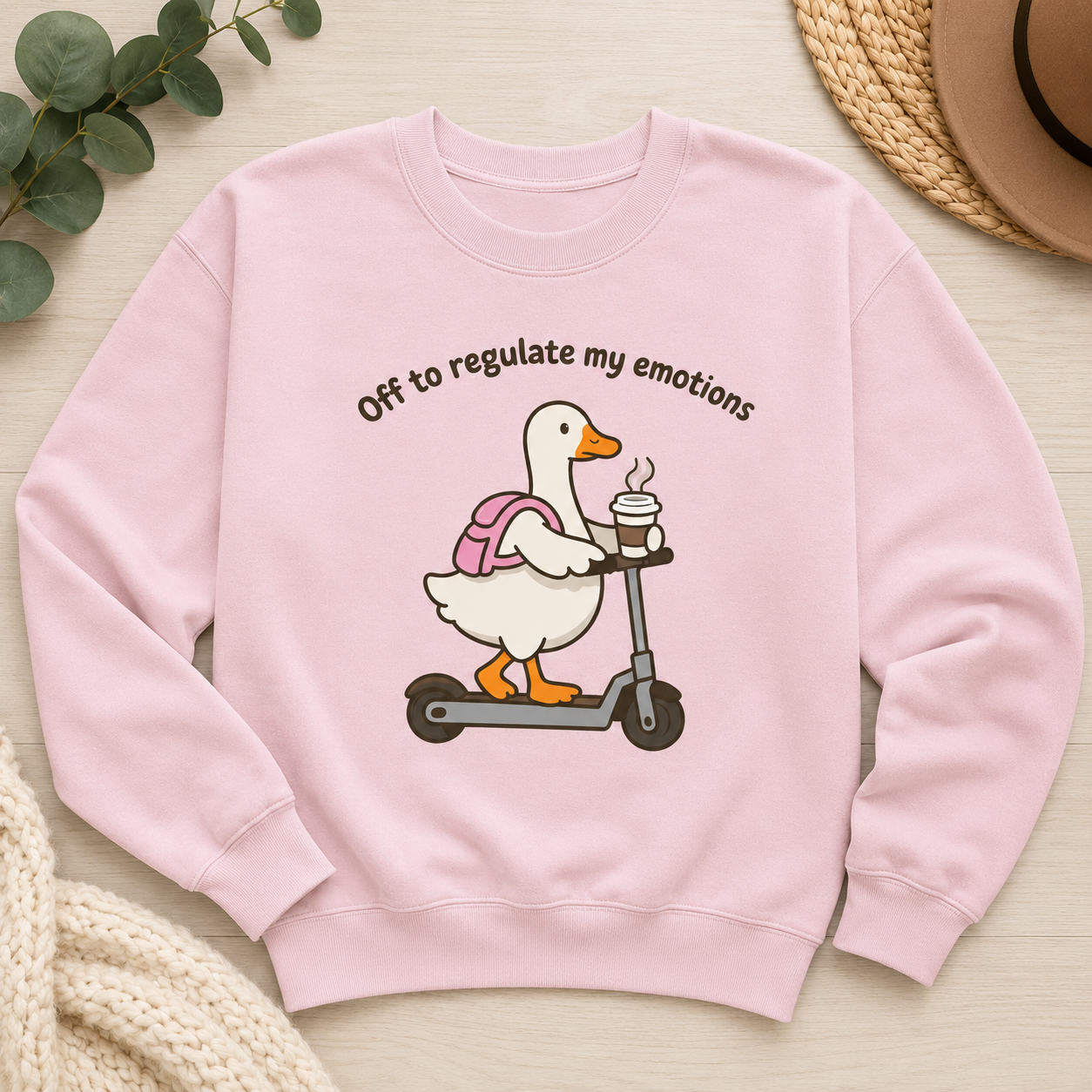 A light pink crewneck sweatshirt laid flat on a light wood surface, featuring a cartoon duck on a scooter with the text 'Off to regulate my emotions.' Decorative props including green leaves, a woven hat, and a knitted blanket are visible around the sweatshirt.