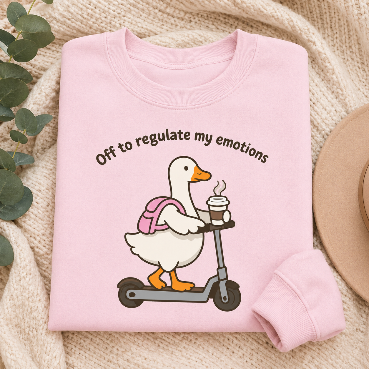 A folded light pink sweatshirt laid flat on a beige knit blanket, with a printed graphic of a white duck on a scooter wearing a pink backpack and holding a coffee cup. The text above reads, 'Off to regulate my emotions.