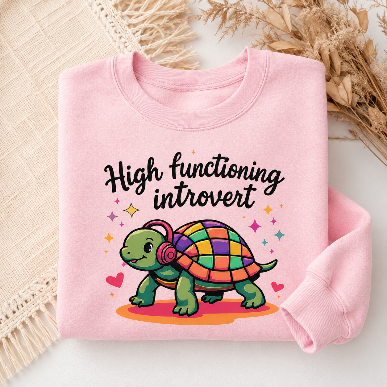 A folded light pink sweatshirt with a graphic print reading 'High functioning introvert' above a colorful cartoon turtle wearing headphones, styled on a neutral surface with dried flowers and a woven textile.