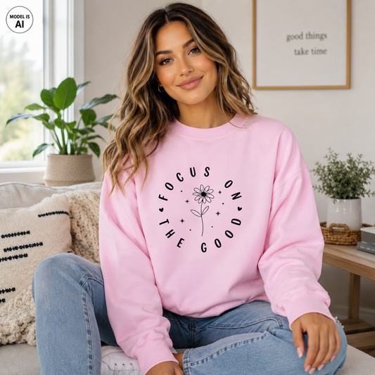 A woman sitting on a couch wearing a light pink crewneck sweatshirt with a black graphic that reads 'FOCUS ON THE GOOD' arranged in a circle around a flower. The background shows a cozy living room setting with a window, houseplant, and framed wall art.