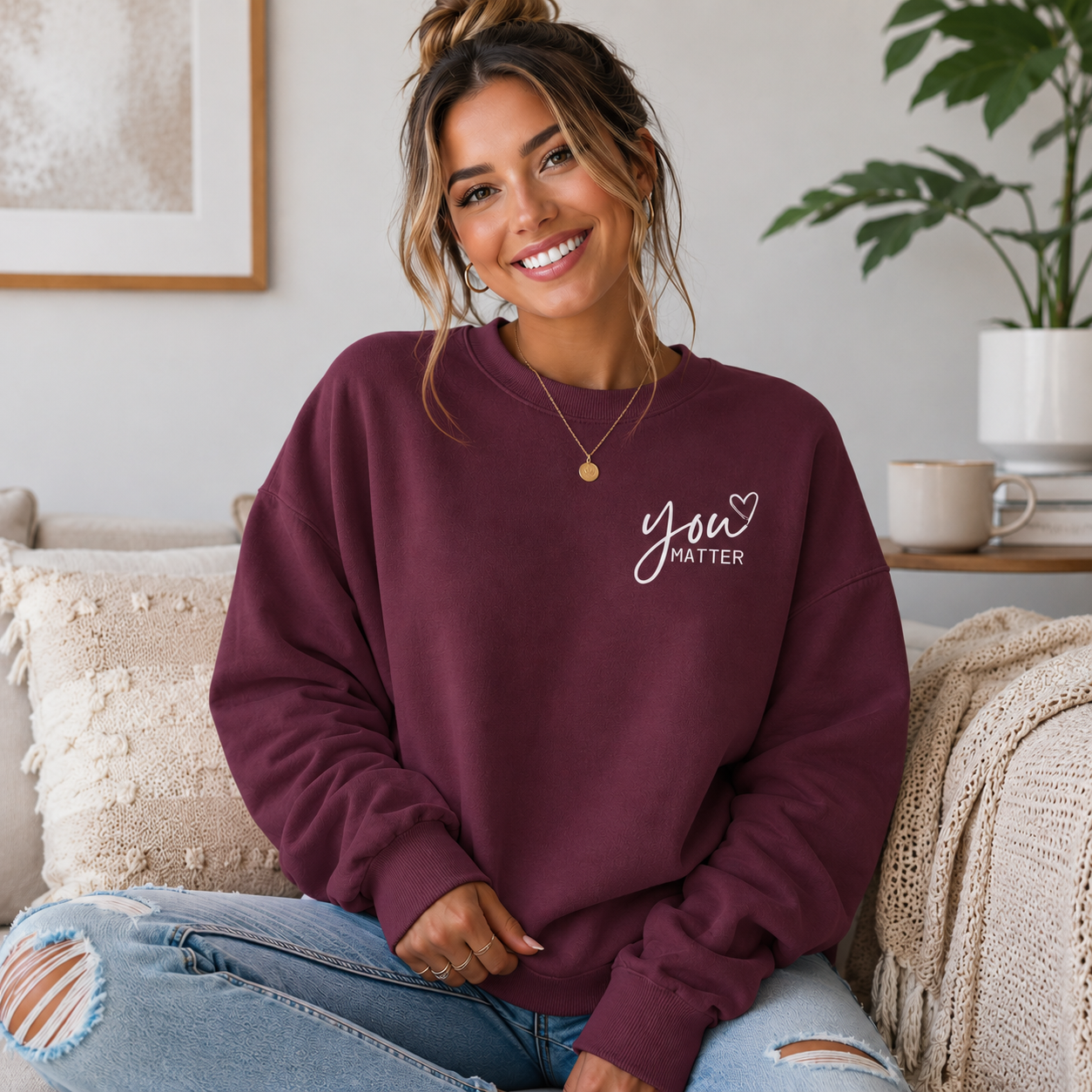 A woman wearing a maroon crewneck sweatshirt with 'You Matter' printed in white script on the chest, sitting on a cozy couch in a bright living room.