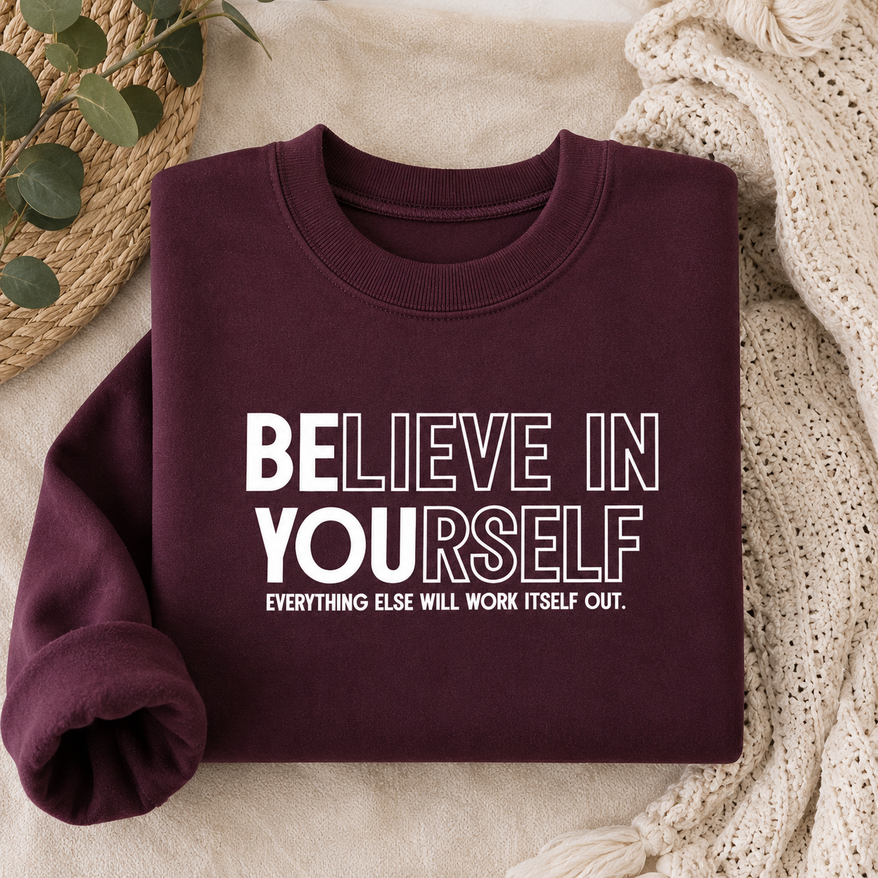 A folded crewneck sweatshirt in deep burgundy with white text reading 'BELIEVE IN YOURSELF' and 'EVERYTHING ELSE WILL WORK ITSELF OUT.' It is styled on a beige fabric surface with a knit blanket, a woven mat, and green leaves.