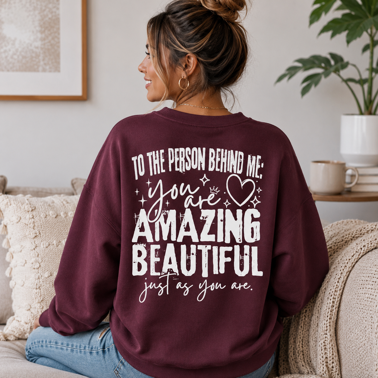 A woman sitting on a couch wearing a maroon sweatshirt with a white graphic text print that reads 'TO THE PERSON BEHIND ME: you are AMAZING BEAUTIFUL just as you are.'.