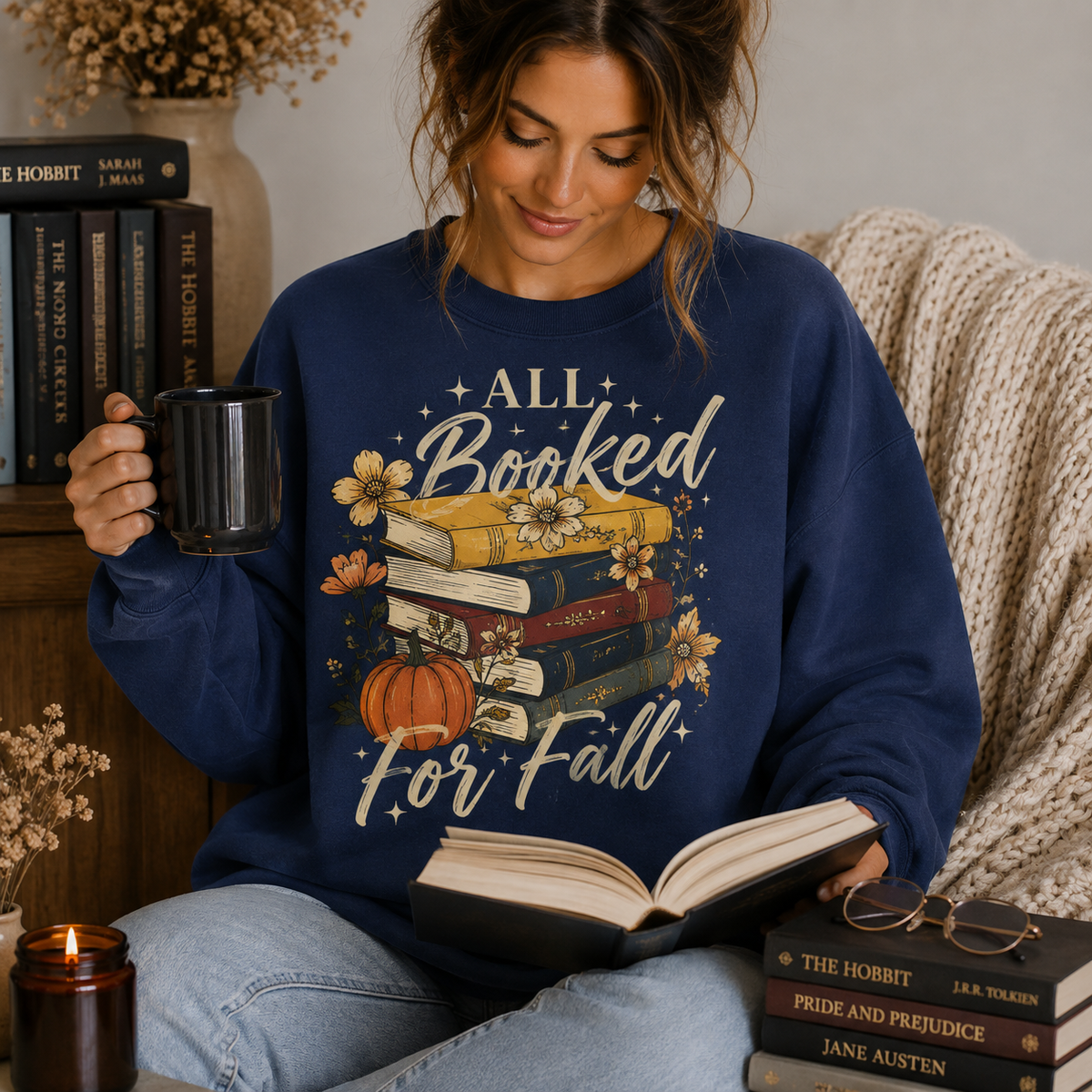 Woman wearing a Navy Fall Book Lover Sweatshirt with All Booked for Fall books and pumpkin graphic while reading and holding a coffee mug.