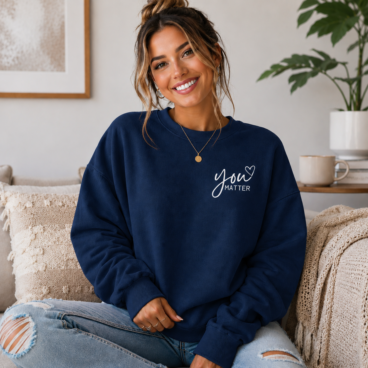 A woman sitting on a cozy couch wearing a navy blue crewneck sweatshirt with 'You Matter' printed in white script on the left chest, paired with distressed light blue jeans.