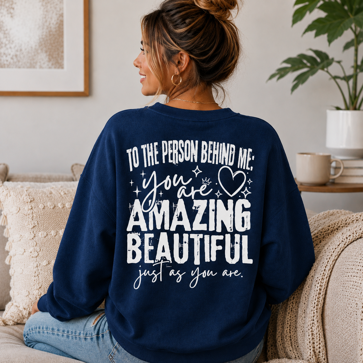 A woman sitting on a couch wearing a navy blue sweatshirt with a white graphic text print that reads 'TO THE PERSON BEHIND ME: you are AMAZING BEAUTIFUL just as you are.'.