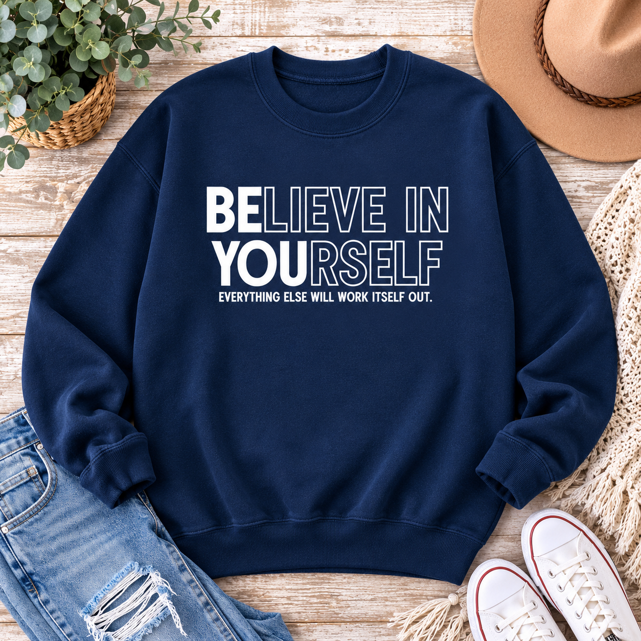 A navy blue crewneck sweatshirt with the motivational text 'BELIEVE IN YOURSELF' in white block letters and 'EVERYTHING ELSE WILL WORK ITSELF OUT.' underneath, styled in a flat lay on a wooden surface with jeans, a hat, shoes, and a plant.