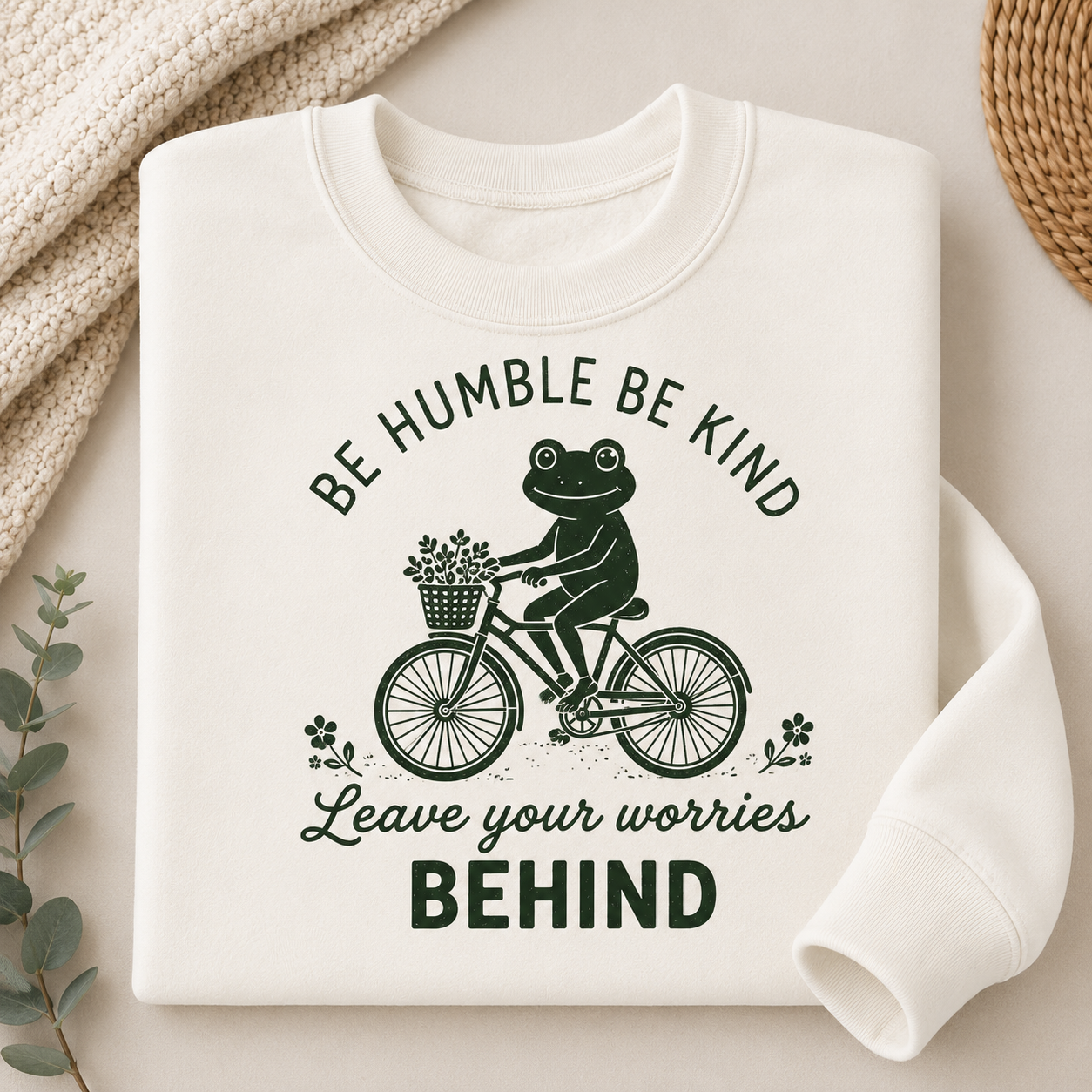 A folded off-white crewneck sweatshirt with a dark green graphic of a frog riding a bicycle and the text 'BE HUMBLE BE KIND Leave your worries BEHIND', styled on a beige surface with a knit blanket, eucalyptus leaves, and a woven mat.