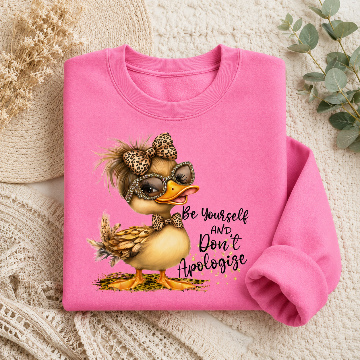 A folded bright pink sweatshirt laid flat on a cream textured fabric background with decorative dried flowers and greenery. The sweatshirt features a cartoon duck wearing leopard-print accessories and sunglasses, with the text 'Be Yourself AND Don't Apologize' printed on the front.