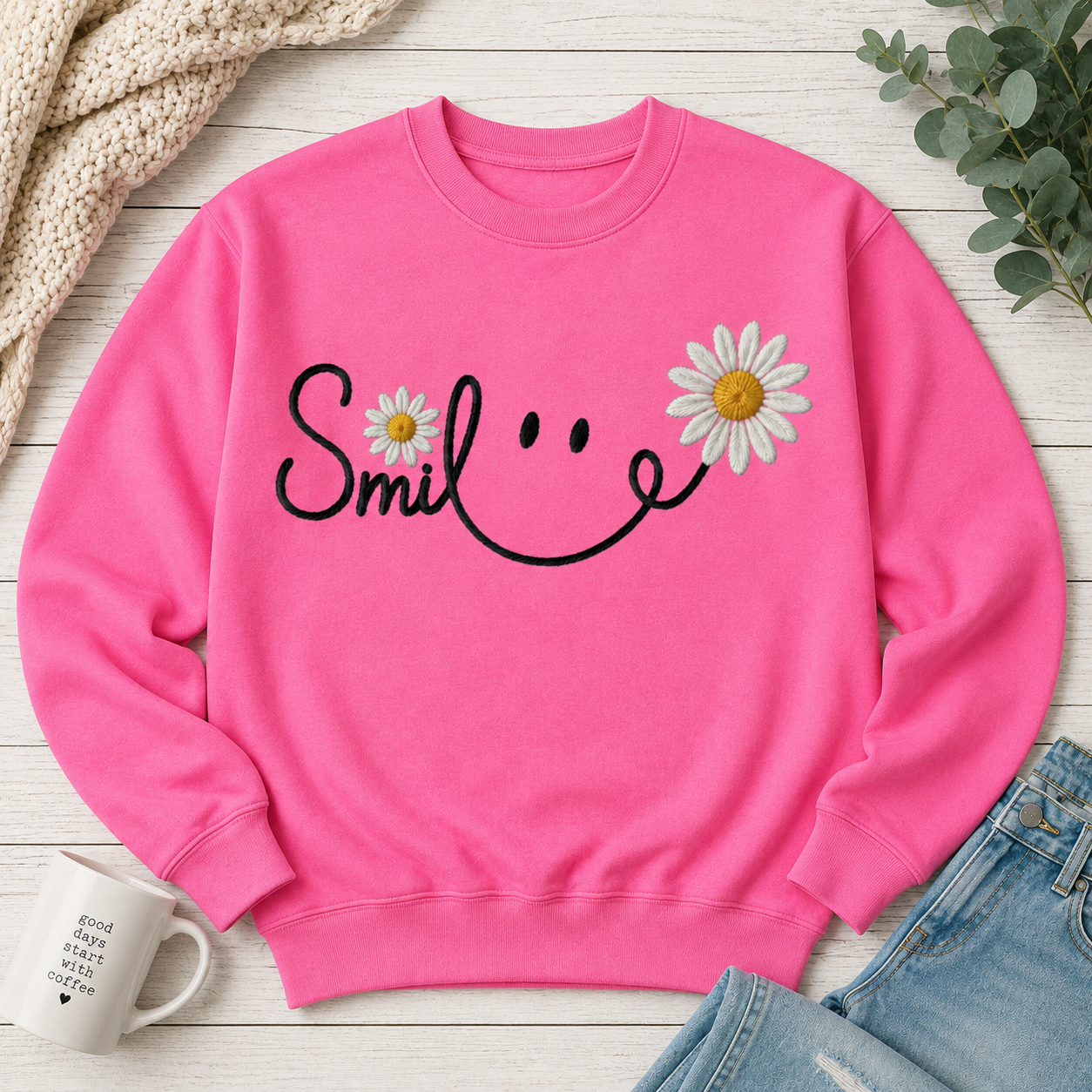 A bright pink crewneck sweatshirt laid flat on a white wood surface, featuring a black smiley face graphic formed by the word 'Smile' with two white daisies with yellow centers. Styled flat-lay props include a chunky knit beige throw, green eucalyptus leaves, blue jeans, and a white ceramic mug.