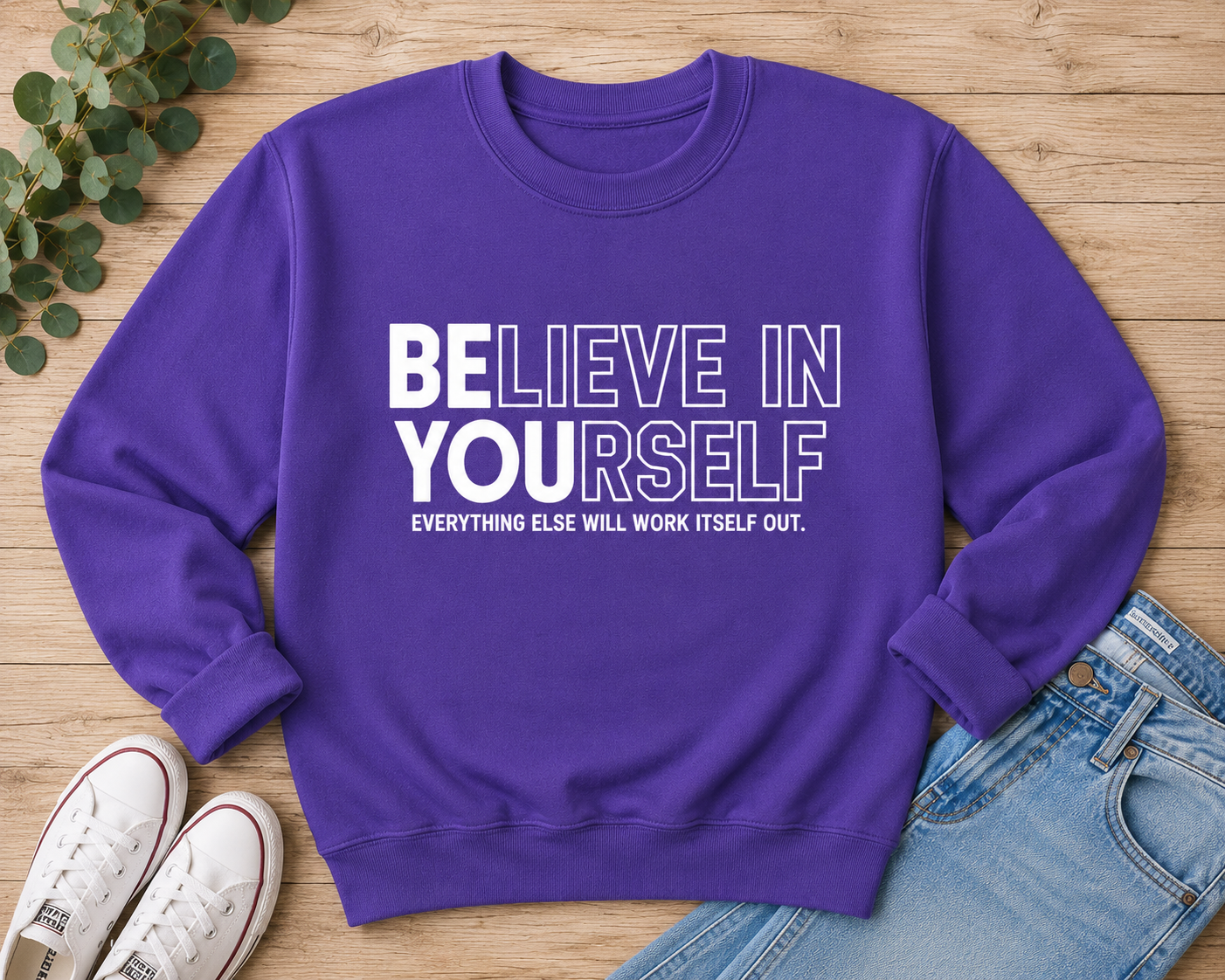 A purple crewneck sweatshirt with white text reading 'BELIEVE IN YOURSELF' and 'EVERYTHING ELSE WILL WORK ITSELF OUT.' laid flat on a wooden surface with jeans, white sneakers, and green leaves as styling props.