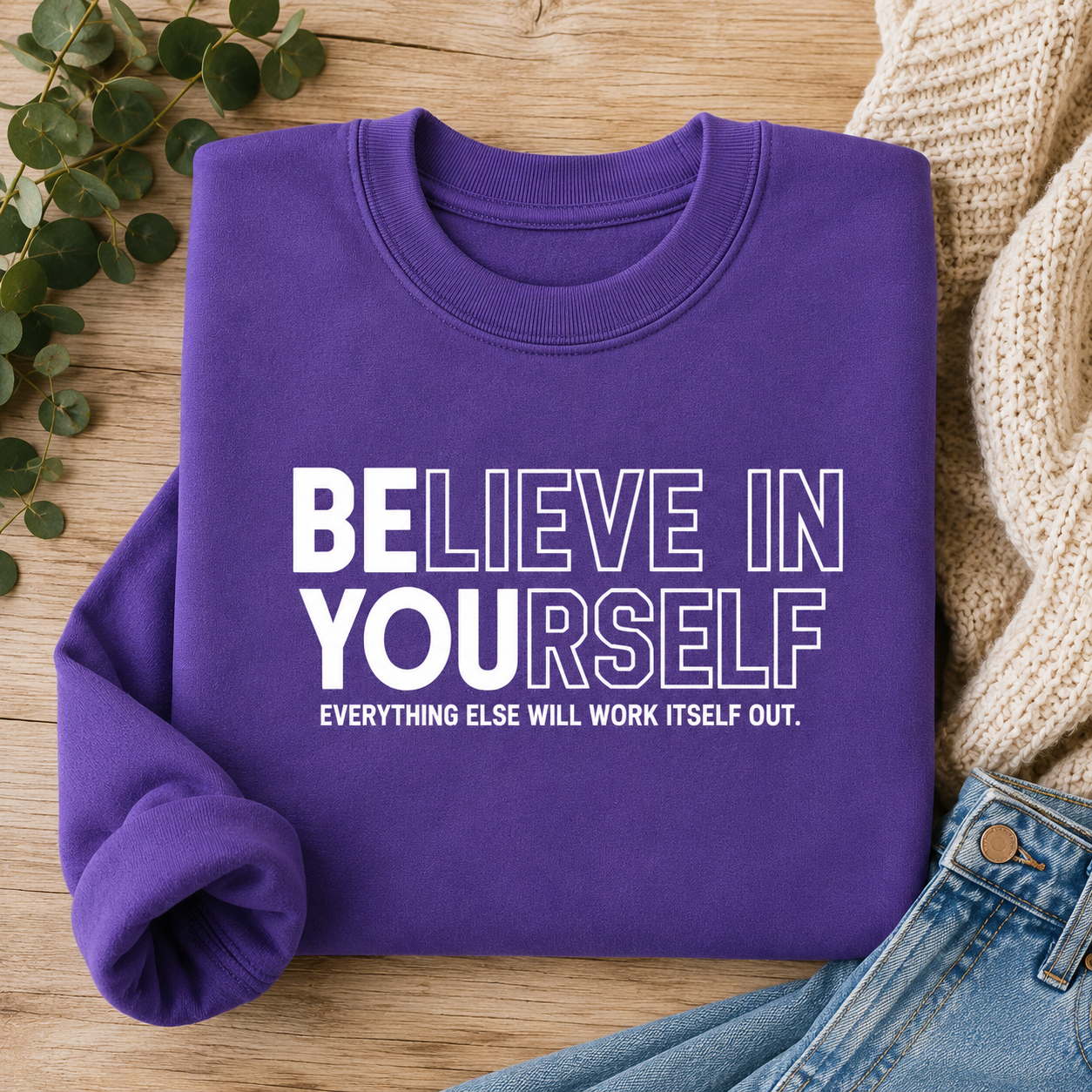 A folded purple sweatshirt with the motivational text 'BELIEVE IN YOURSELF' and 'EVERYTHING ELSE WILL WORK ITSELF OUT.' printed in white on the front, styled on a wooden surface with eucalyptus leaves, a cream knit sweater, and blue jeans.