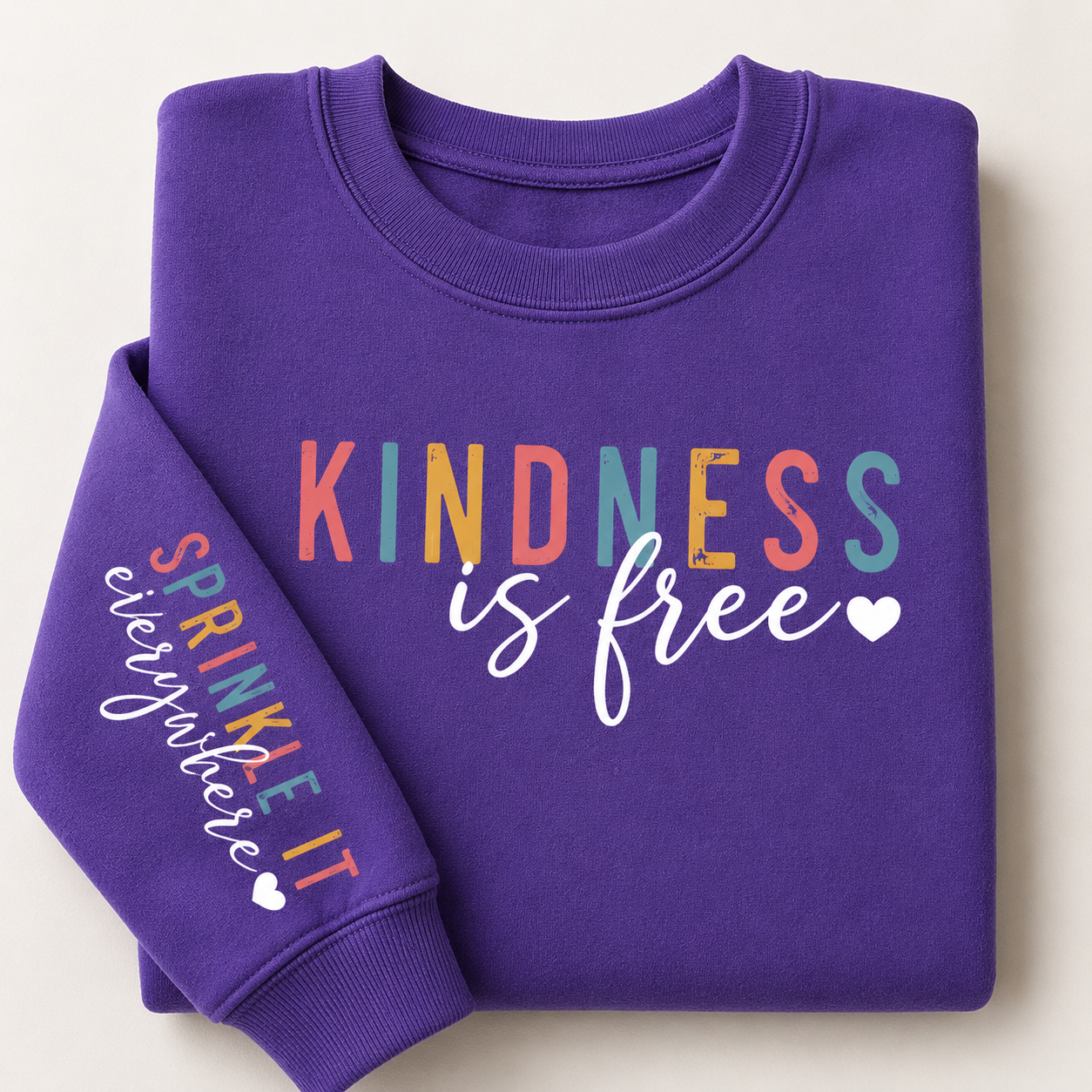 A folded purple crewneck sweatshirt on a light beige background. The front of the sweatshirt features the text 'KINDNESS is free' in multicolor and white script, while the left sleeve displays 'SPRINKLE it everywhere' in multicolor script with a small white heart.