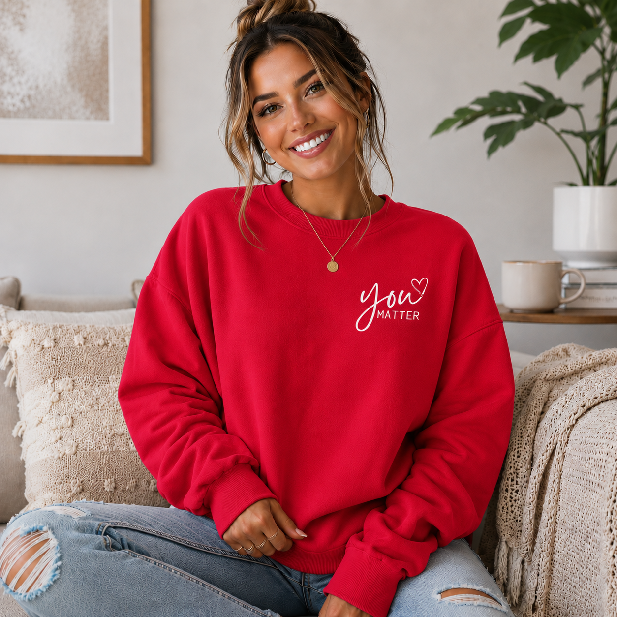 A woman sitting on a couch wearing a bright red crewneck sweatshirt with 'You MATTER' printed in white script on the left chest, paired with distressed light blue jeans.